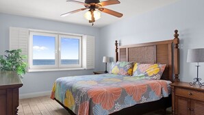 Ein Schlafzimmer mit einem Bett, einem Deckenventilator, einem Fenster mit Rollläden, einem Nachttisch, einer Lampe und einer Pflanze.