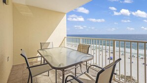A balcony with a glass table and chairs, overlooking a beach with umbrellas and people. - Clearwater Unit 7C (Gulf Shores)