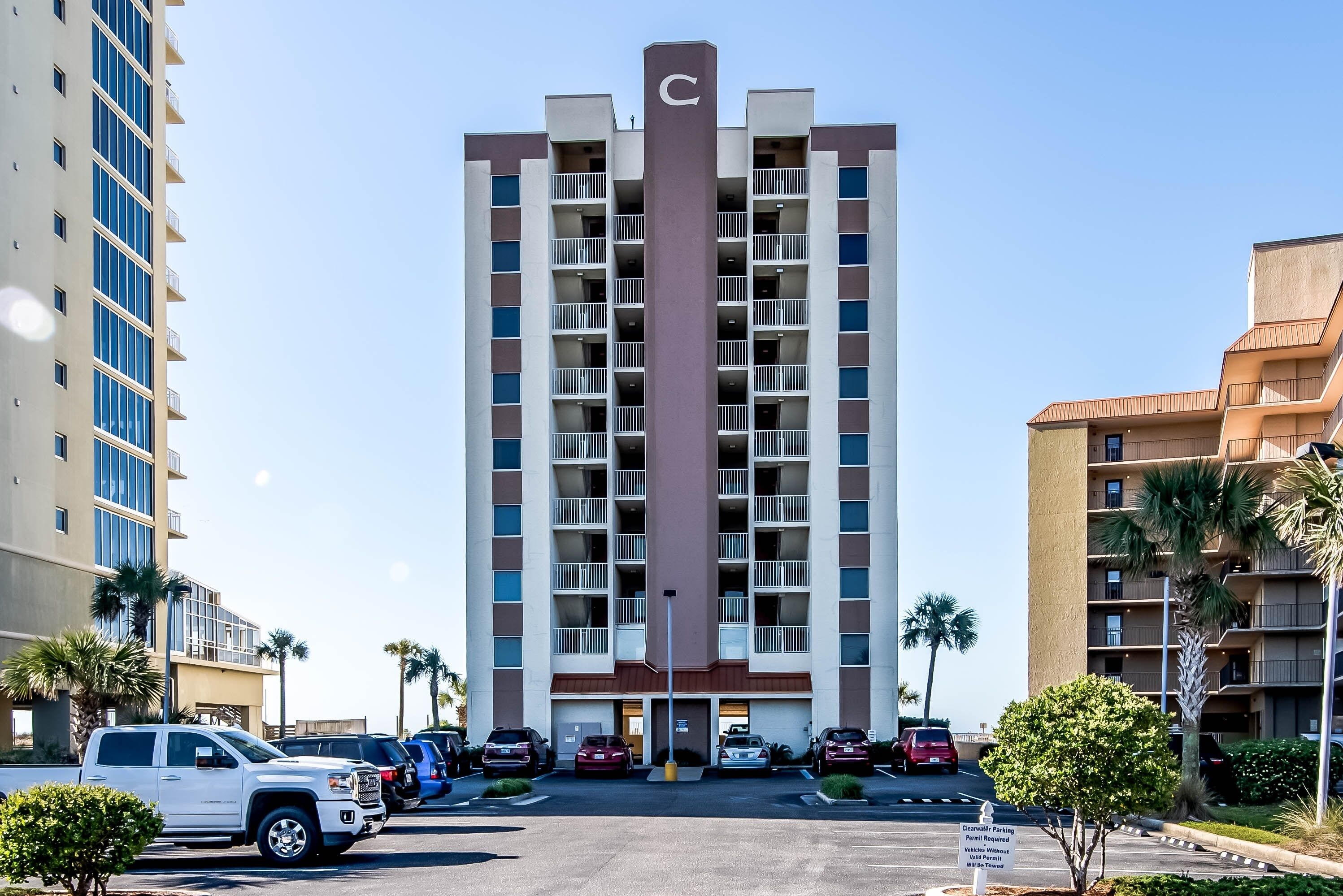 A multi-story hotel with a central vertical feature, parking in front, and palm trees around.