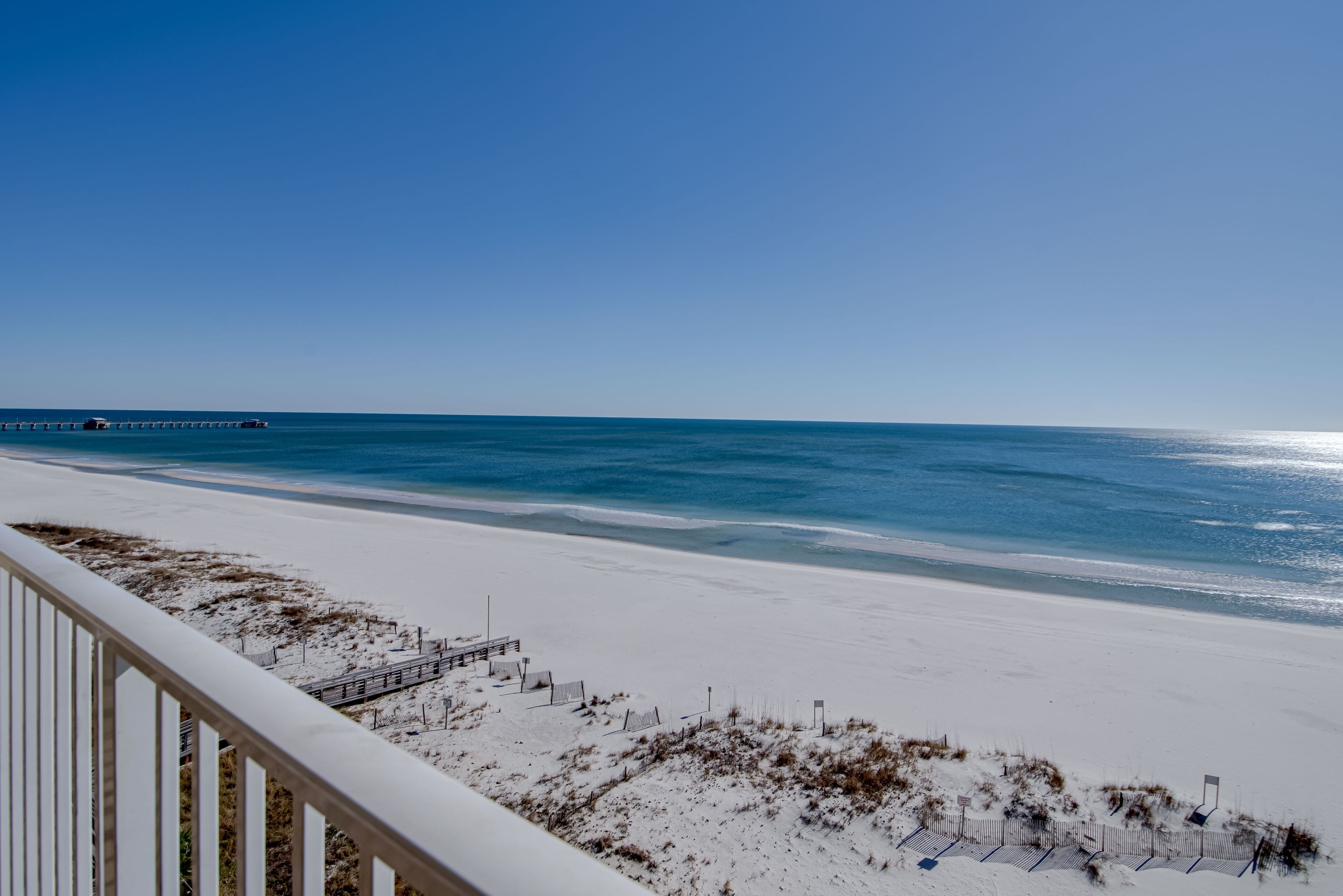 A beach with a wooden walkway leading to the water, a pier in the distance, and a clear blue sky.