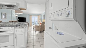 A kitchen with white appliances, a dining area with a chair, and a view through the window. - Clearwater Unit 7C (Gulf Shores)