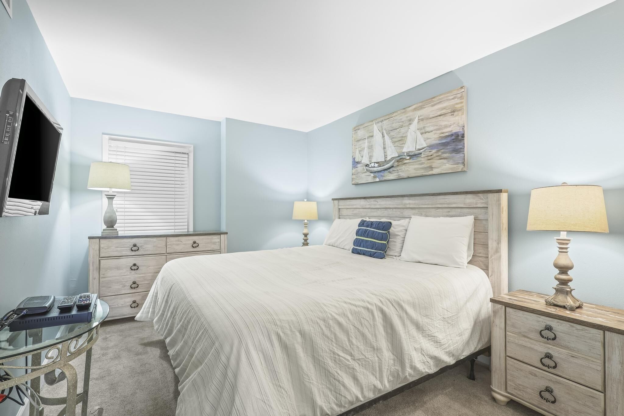 A bedroom with a bed, two bedside tables with lamps, a dresser, a TV, and a window with blinds.
