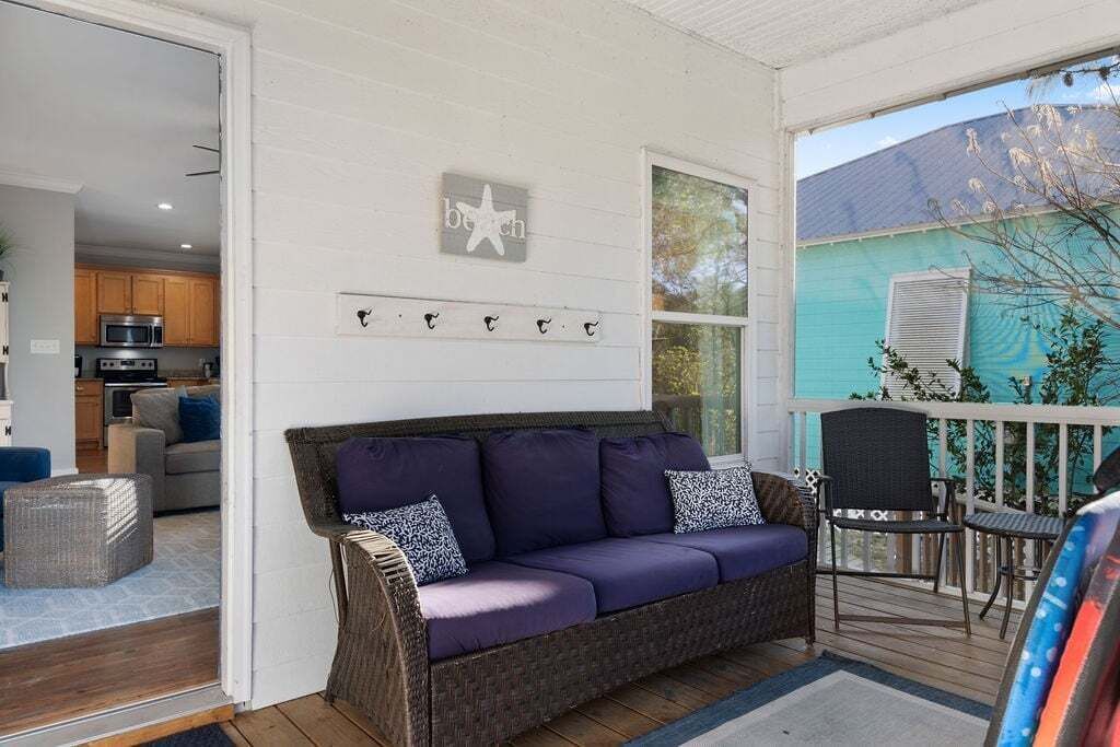 A cozy living area with a purple sofa, wicker chairs, and a small table. A kitchen is visible in the background, and a teal house is seen through the window.