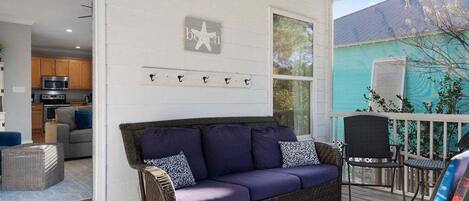 A cozy living area with a purple sofa, wicker chairs, and a small table. A kitchen is visible in the background, and a teal house is seen through the window.
