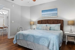 A bedroom with a bed, bedside lamps, a painting of the ocean, a chair, and a desk.