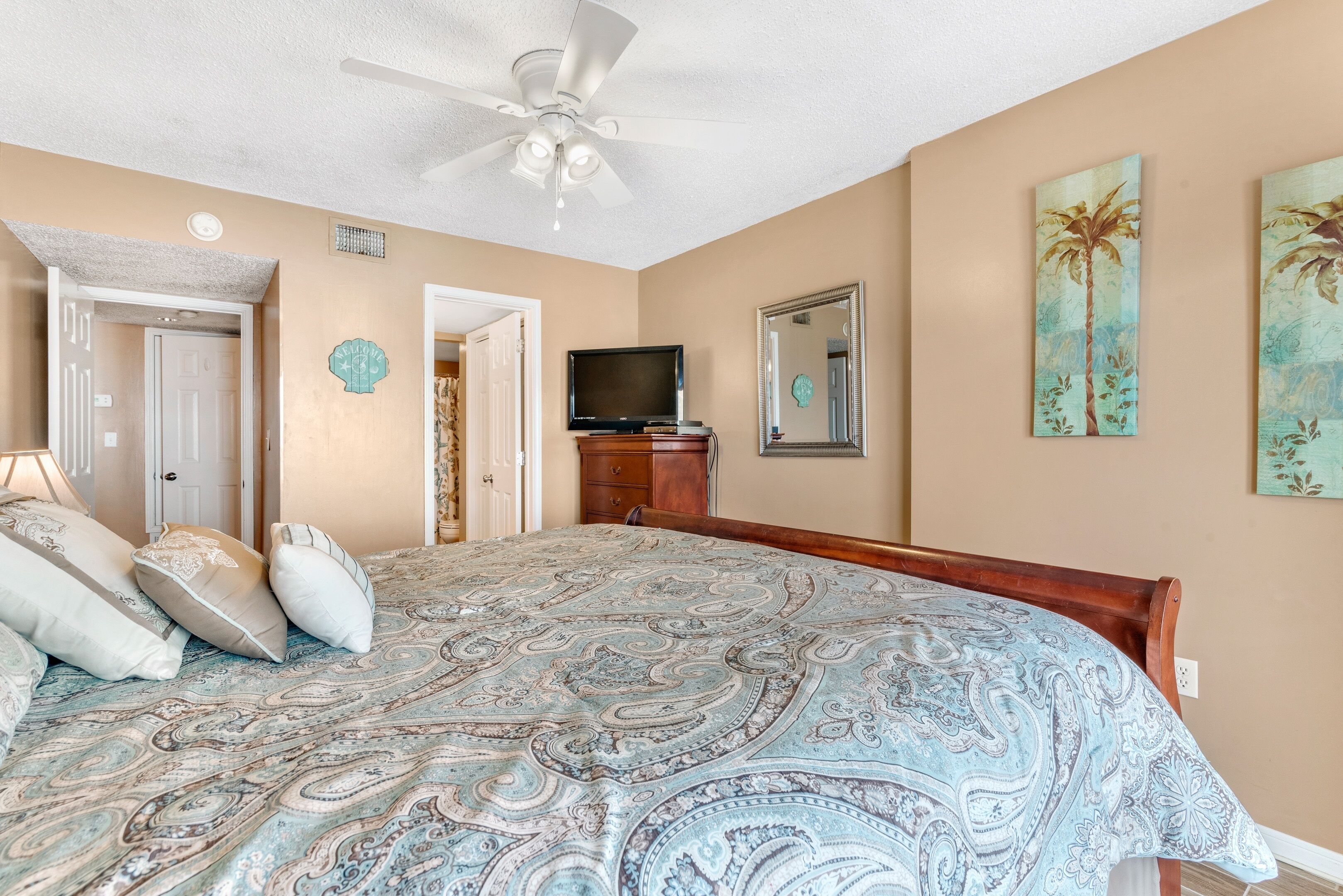 A bedroom with a large bed, a ceiling fan, a television, and artwork on the walls.