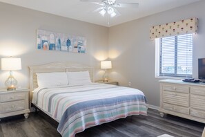 A bedroom with a bed, bedside lamps, a nightstand, a dresser, a ceiling fan, a window with blinds, and a wall-mounted artwork. - Tropical Winds Unit 301 (Gulf Shores)