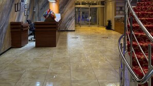 Un vestíbulo de hotel con una recepción de mármol, una escalera con alfombra roja y un amplio ventanal con vitrales.
