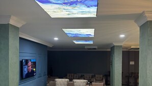 Amplia sala de estar con asientos cómodos, un televisor de pantalla plana y pantallas en el techo que muestran arte abstracto.