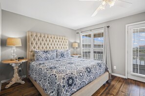 A bedroom with a tufted headboard, a patterned bedspread, two bedside tables with lamps, and a view through a window with blinds. - 214 Shoreland (Gulf Shores)