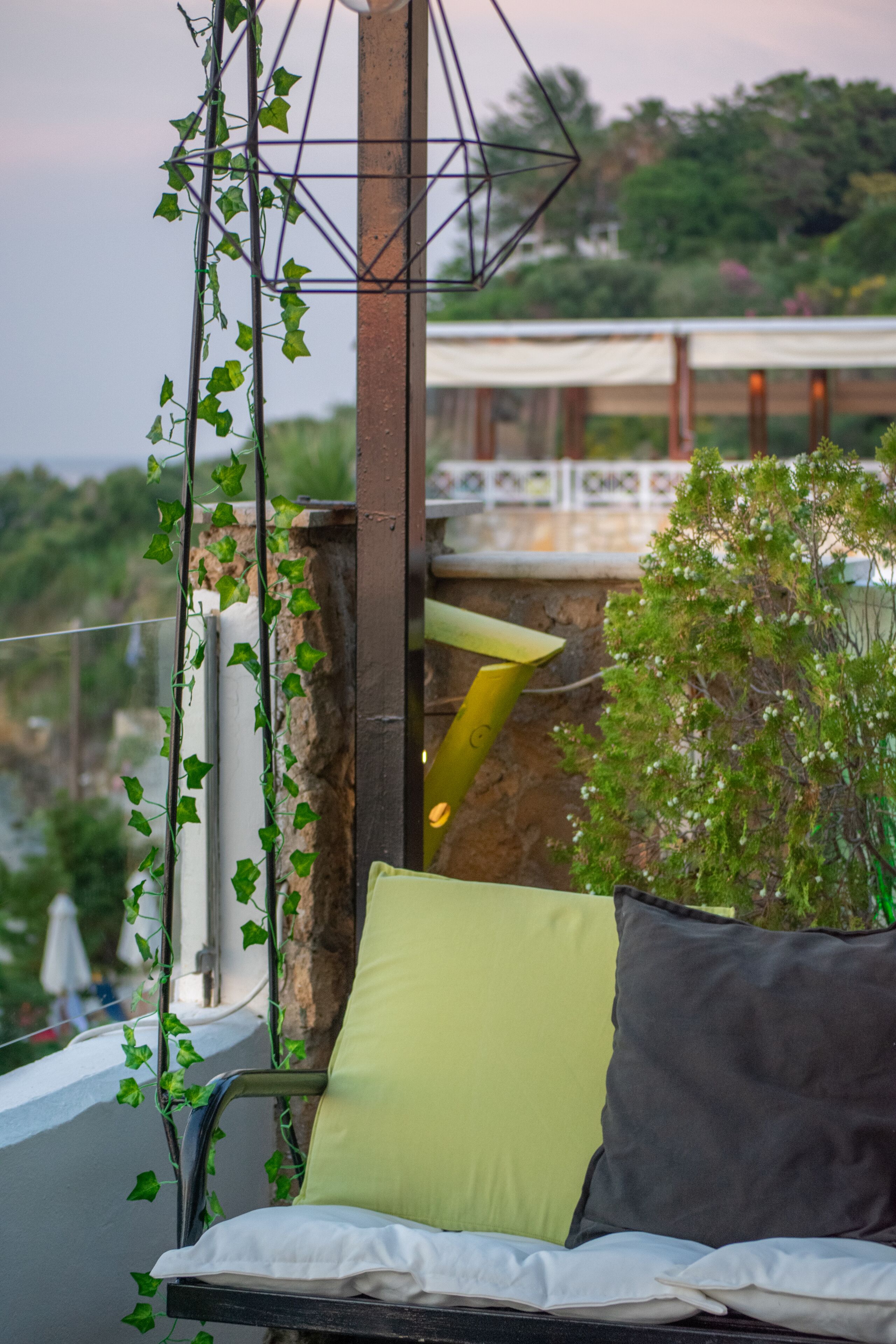 A balcony with a metal chair and cushions, surrounded by greenery and a view of a building in the background.