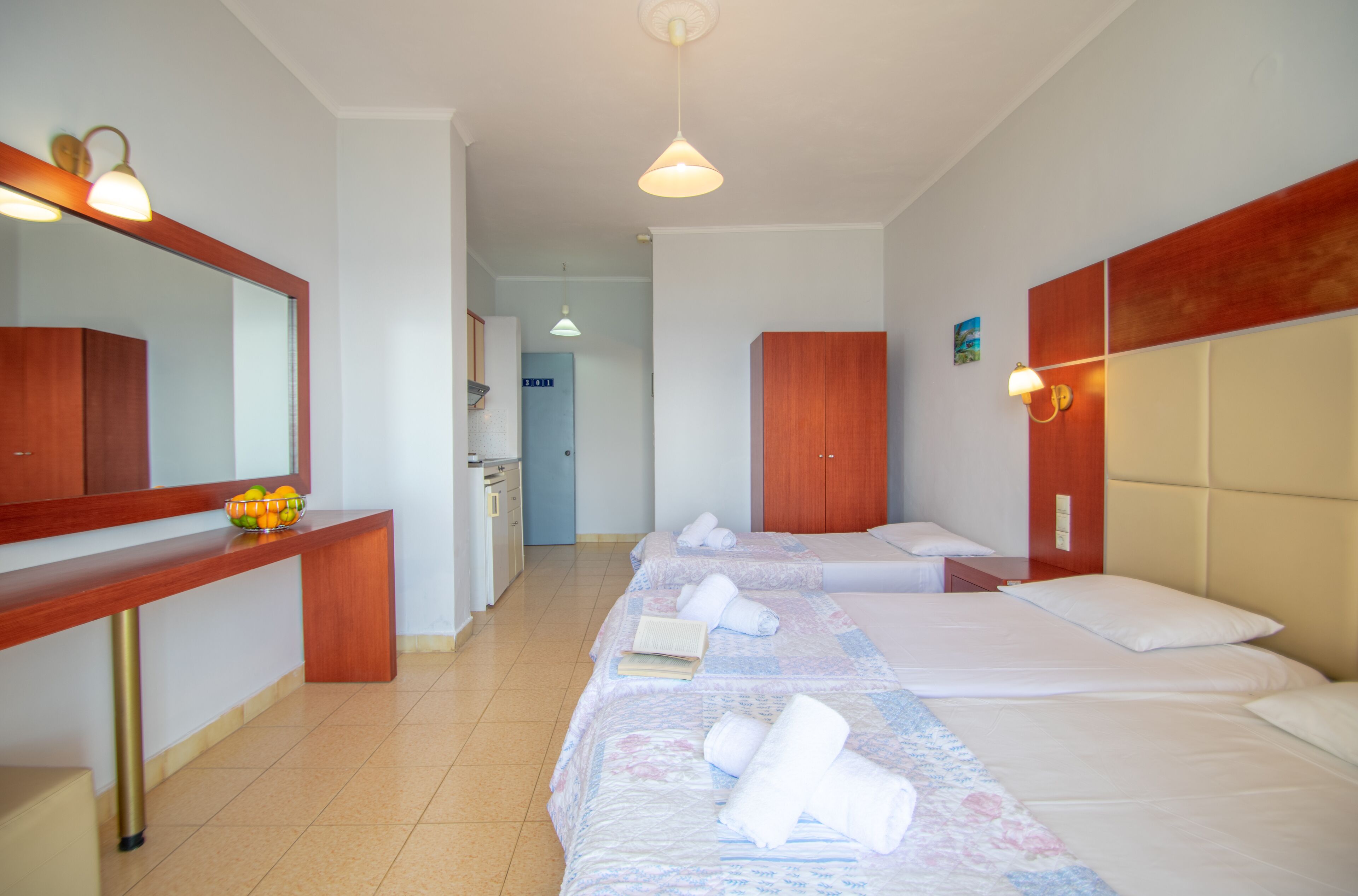 Ein Hotelzimmer mit drei Betten, einem Holztisch, einem Spiegel und einem Badezimmer im Hintergrund.