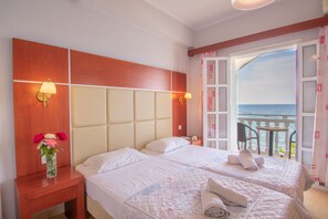 A hotel room with two beds, a nightstand with a vase of flowers, and a balcony with a view of the sea.