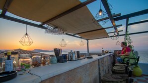 Zona de bar con luces colgantes, una barra con varios artículos y vista al mar al atardecer.