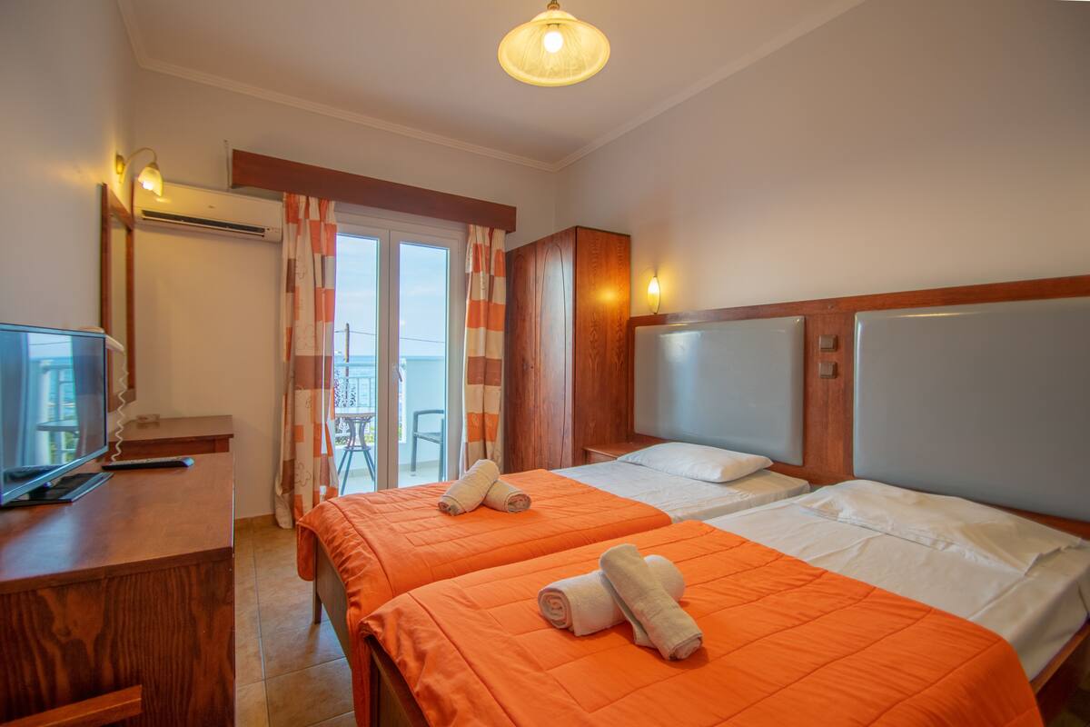 A hotel room with two beds, a desk with a television, and a balcony with curtains.
