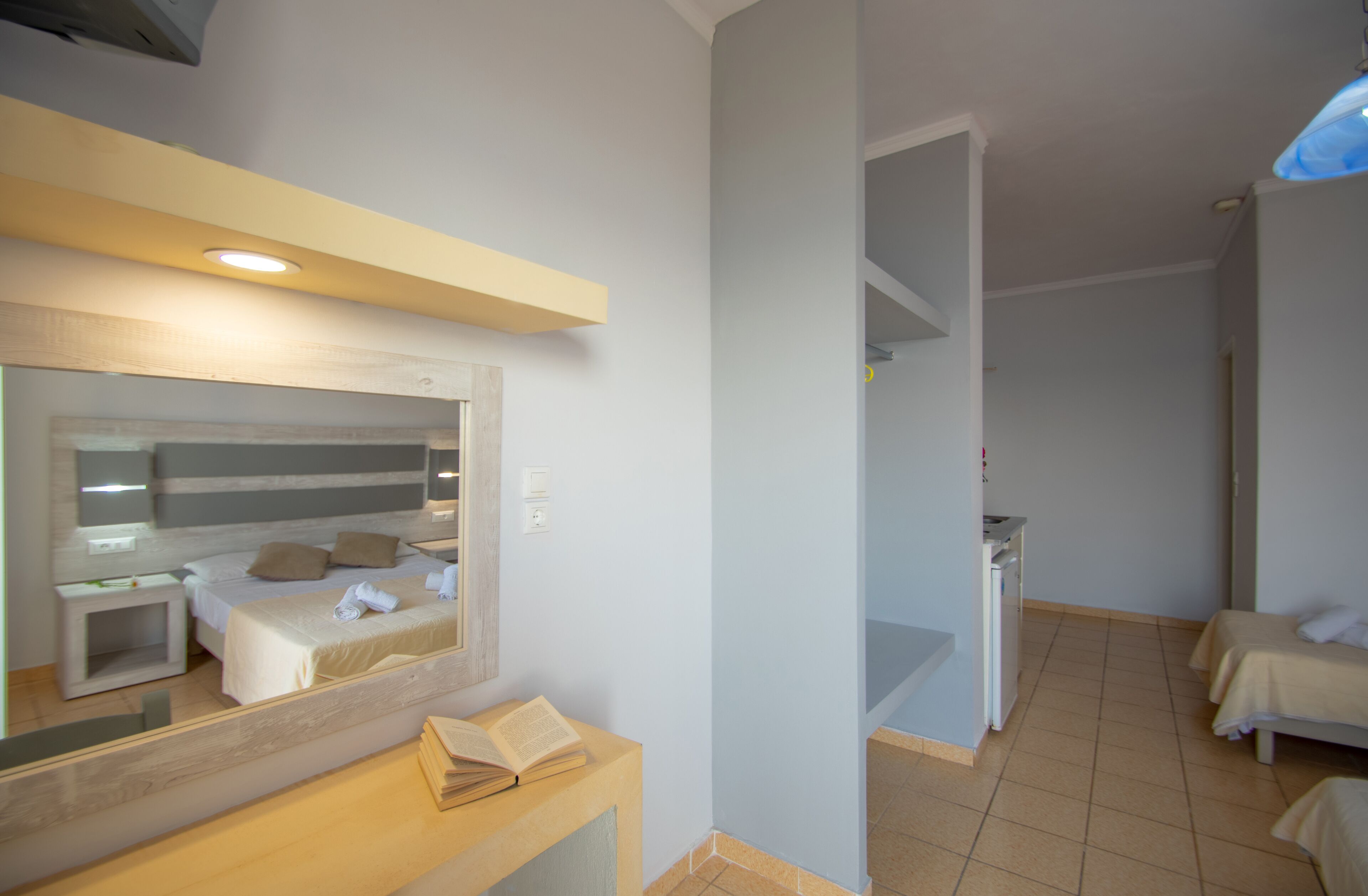 Habitación de hotel con cama, mesita de noche y espejo.  Incluye un escritorio pequeño con una silla y una lámpara. El piso es de baldosas y hay una puerta que da a otra habitación.