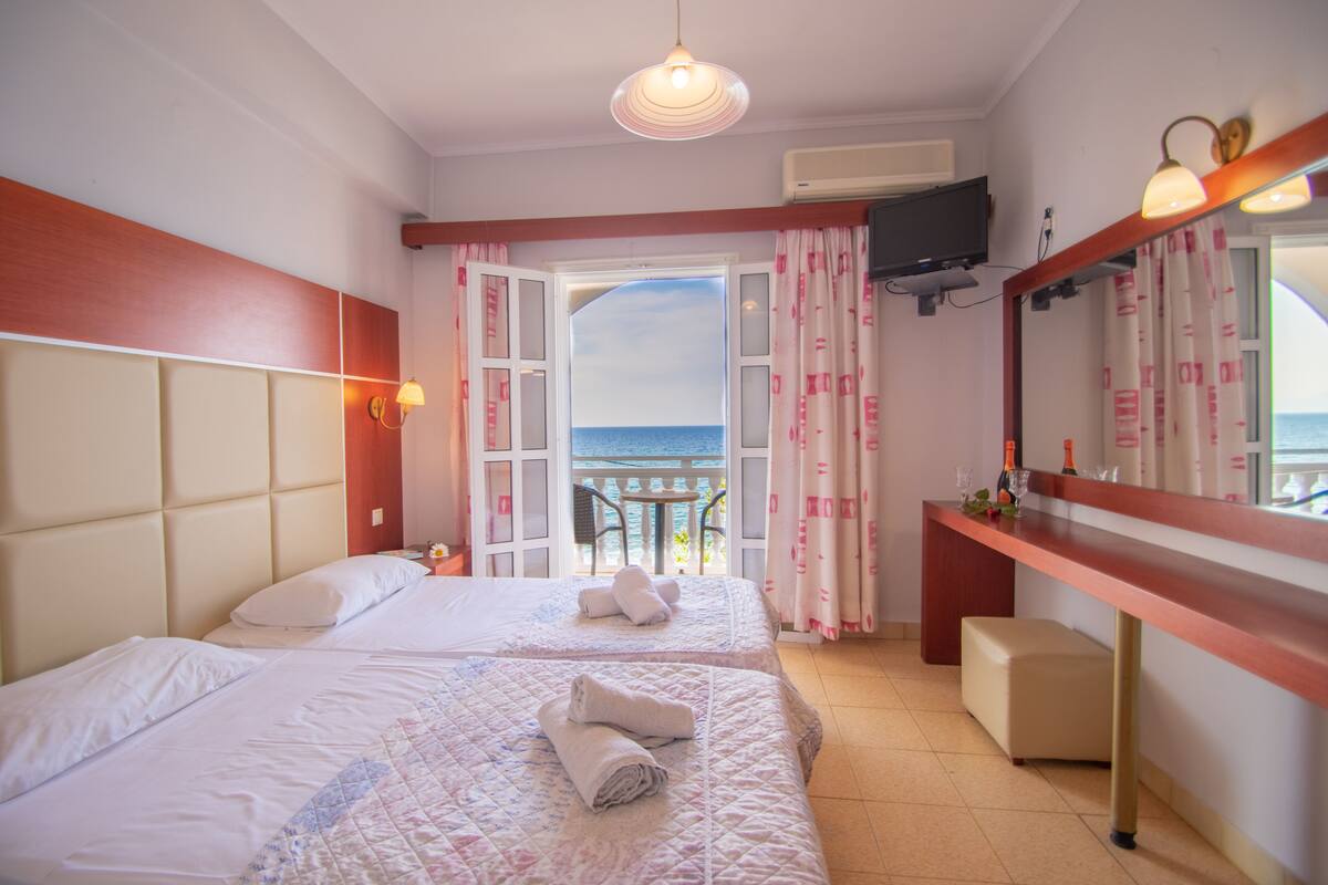 A hotel room with two beds, a balcony with a view of the sea, a TV mounted on the wall, and a small table with a mirror.