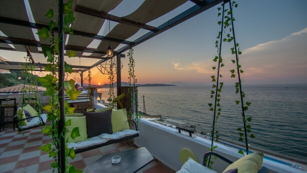 Una terraza con suelo a cuadros, mobiliario exterior con cojines, plantas colgantes y vistas al mar al atardecer.