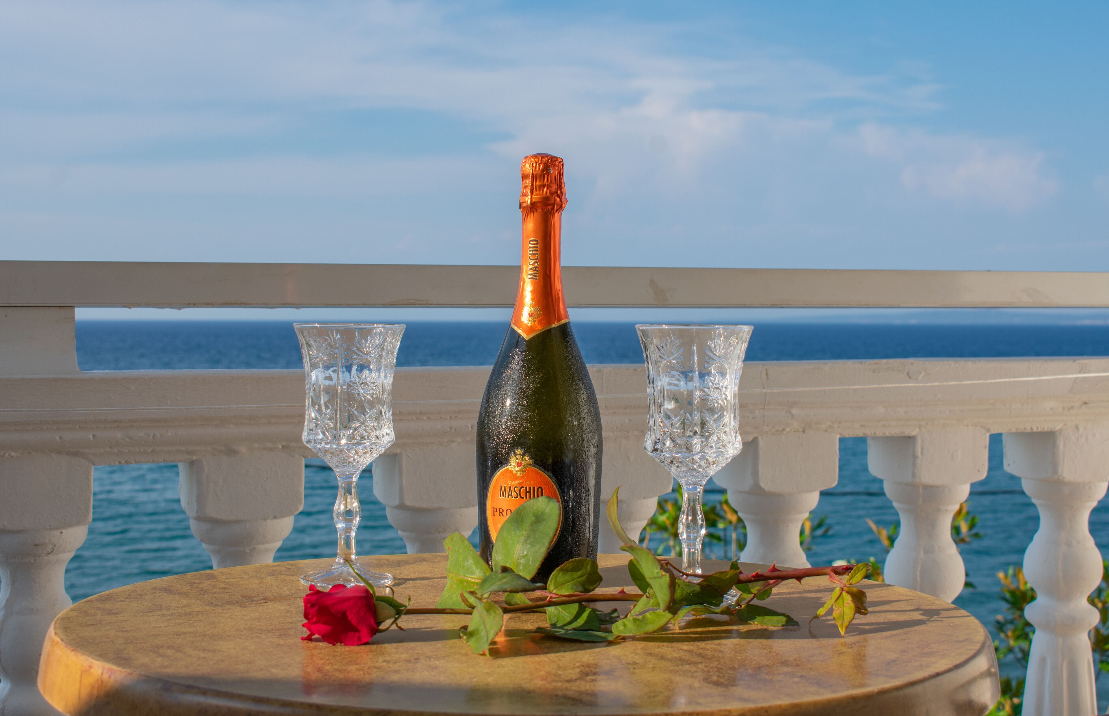 A bottle of Maschio Prosecco on a round table with two glasses and a rose, set on a balcony with a view of the sea.