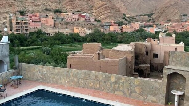 A swimming pool with a stone wall and a view of a village with traditional buildings and mountains in the background. -  Les Jardins de Todgha (Toudgha El Oulia)