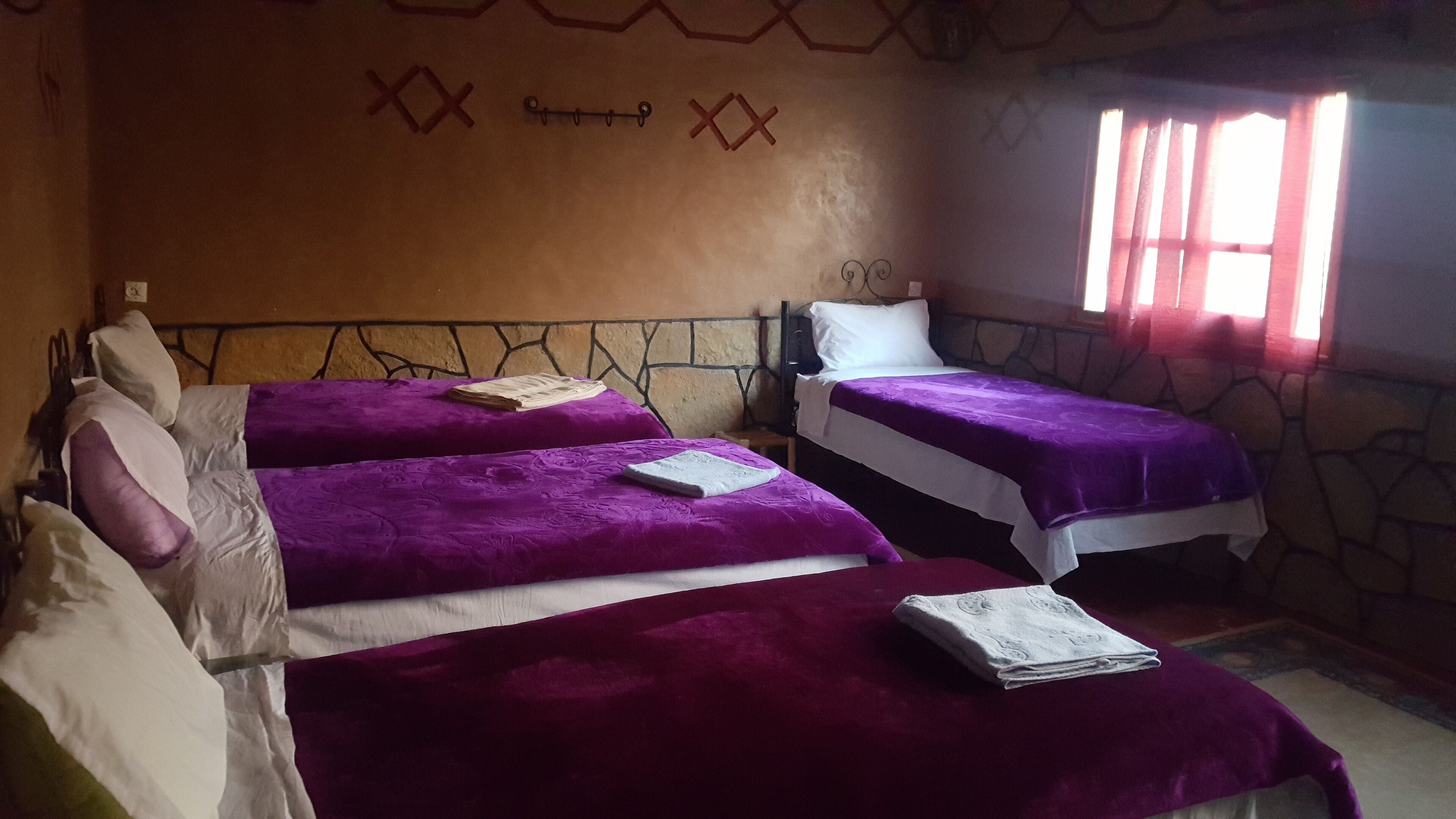 A room with two beds, each with a purple bedspread and white pillows. There is a stone wall with a decorative pattern and a window with red curtains.