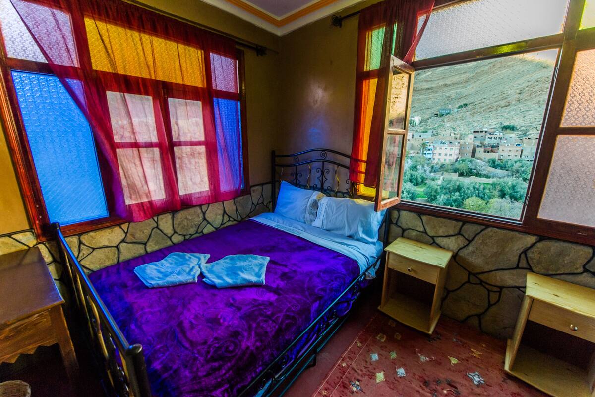 A room with a bed, a window with a view of a town, and a stone wall.