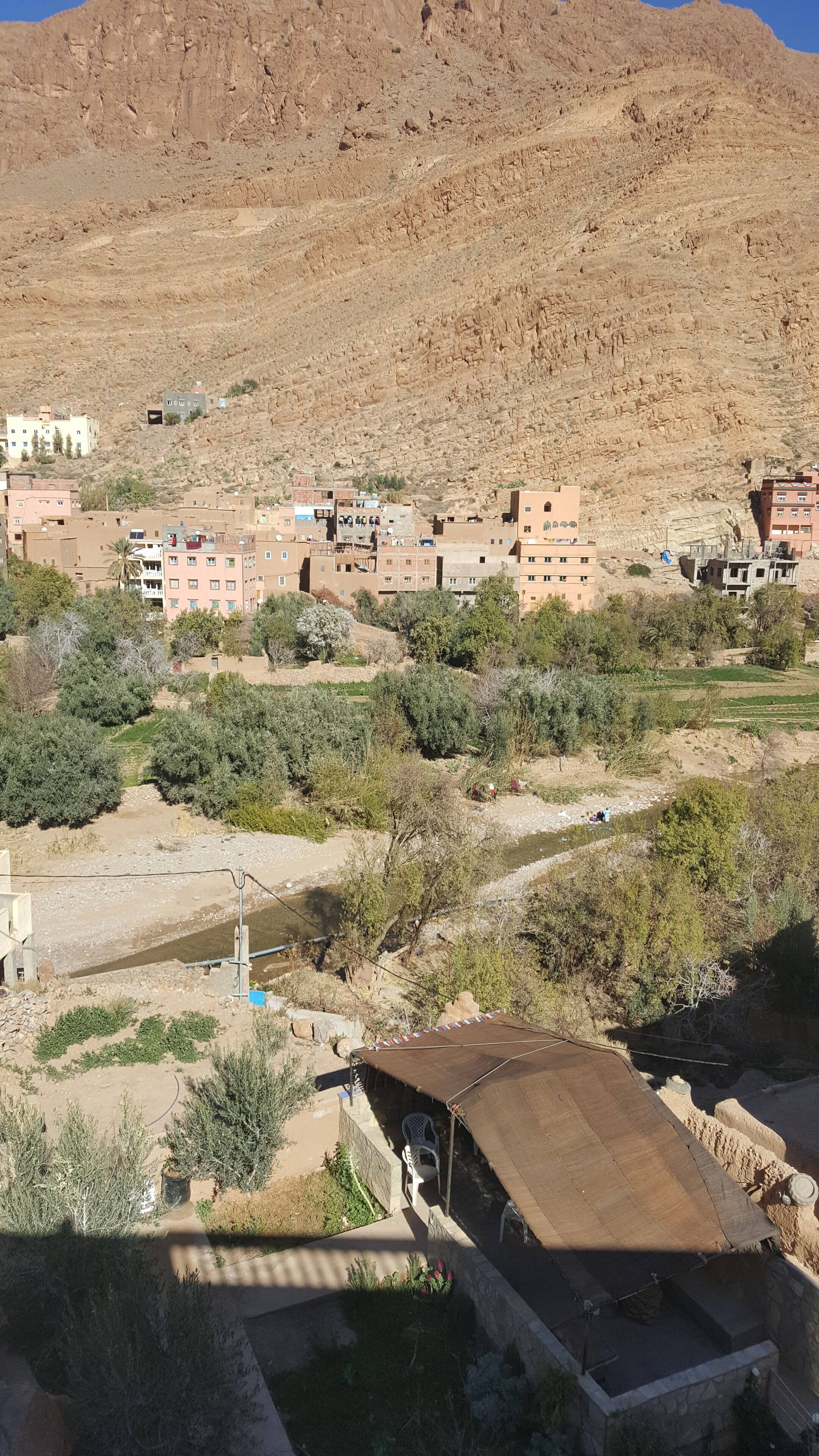 A residential area with houses, some with flat roofs and others with pitched roofs, surrounded by sparse vegetation and a hill in the background.