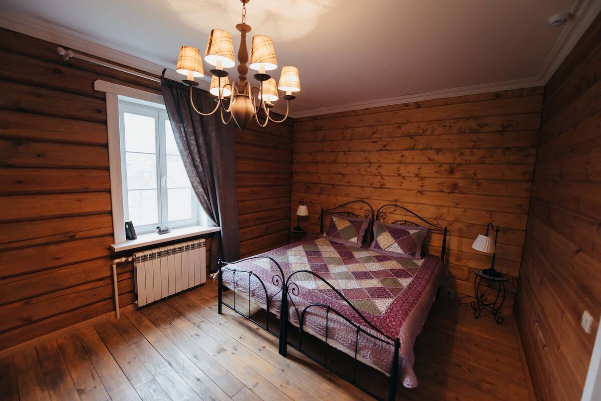 Ein Zimmer mit Holzwänden, einem Bett, einem Fenster mit Vorhängen, einem Heizkörper und einer Chandelüstere.