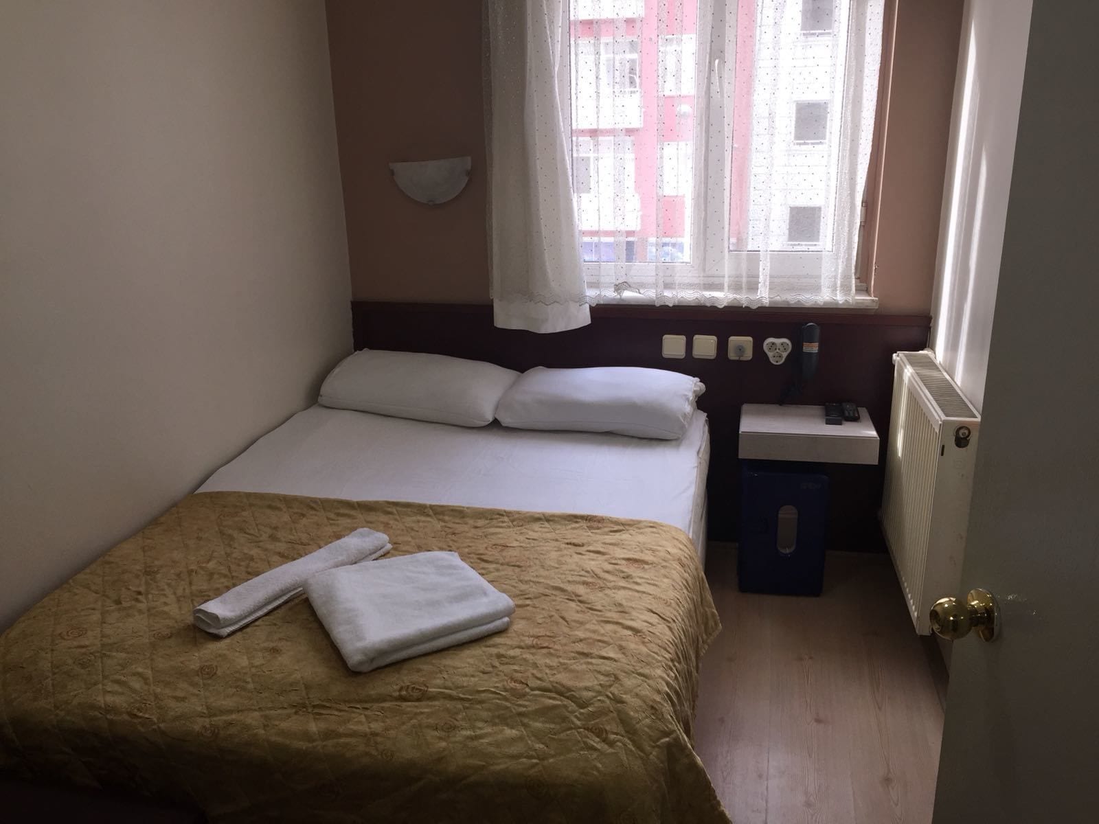 Een kleine, verzorgde slaapkamer met een enkel bed, een nachtkastje, een radiator en een raam met uitzicht op gebouwen.