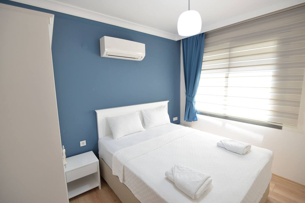 Habitació amb un llit, roba de llit blanca, una taula de nit, un aparell d'aire condicionat, una finestra amb cortines i una làmpada al sostre.