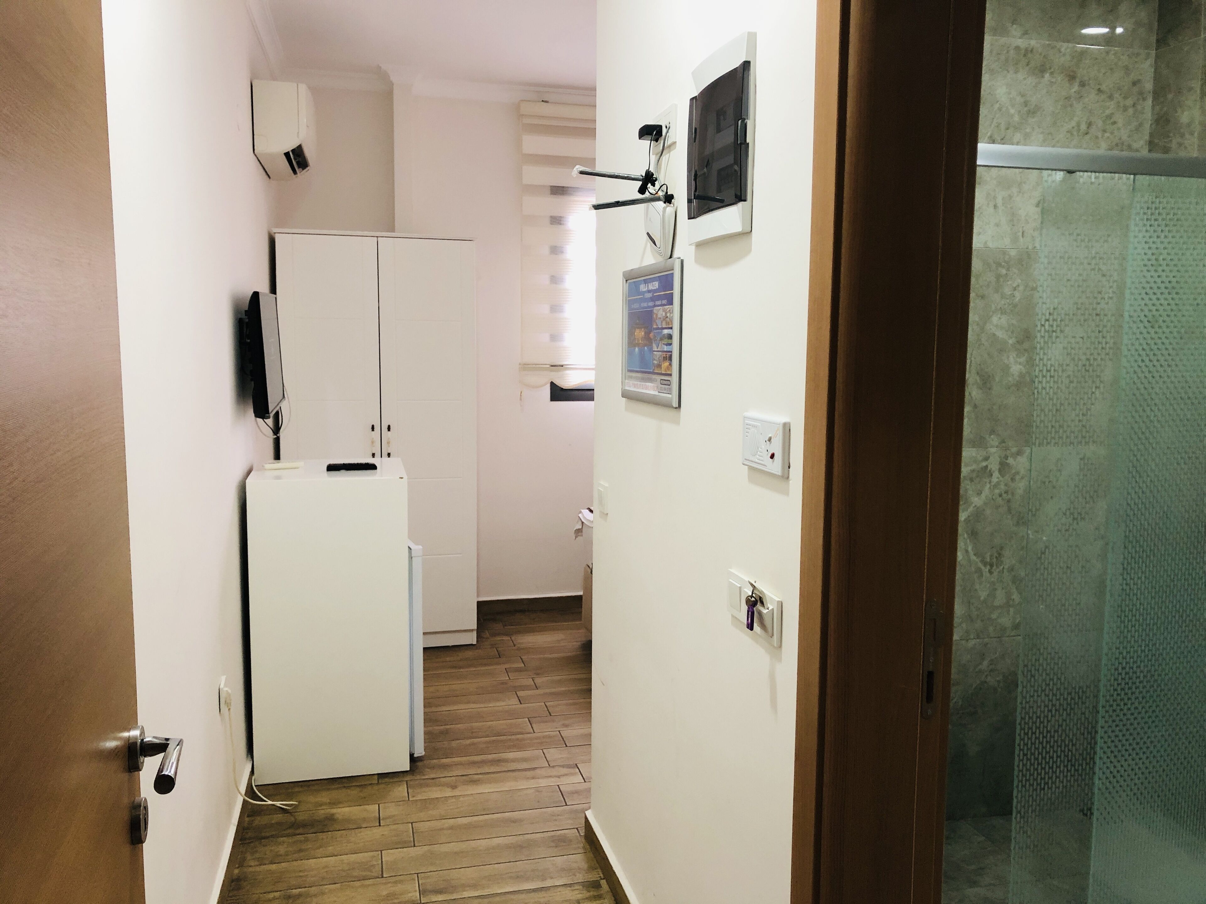 Un couloir étroit avec un réfrigérateur blanc, un climatiseur fixé au mur et une porte dotée d’un cadre en bois.