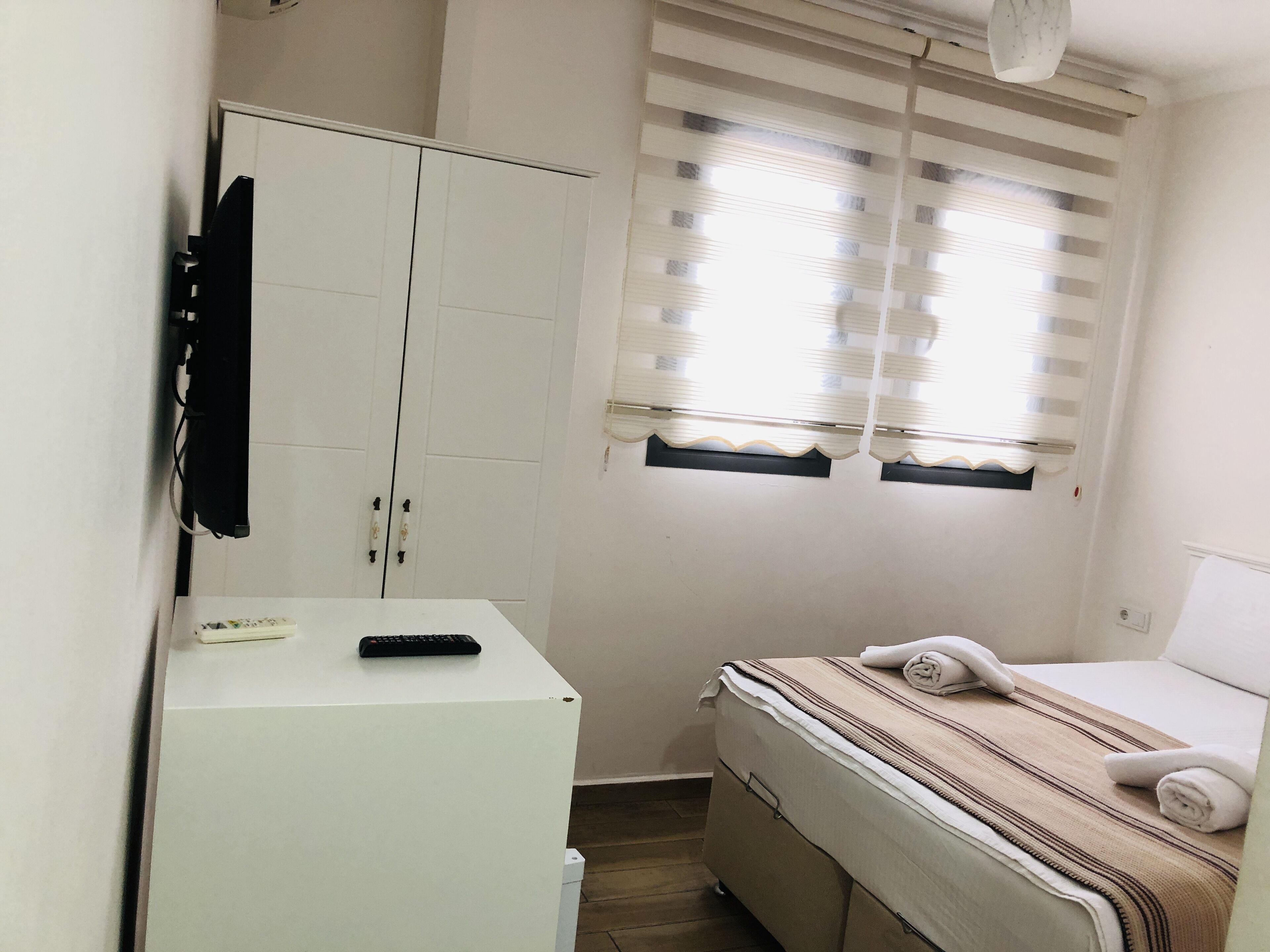 A small, clean room with a bed, a white wardrobe, and a desk with a remote control.