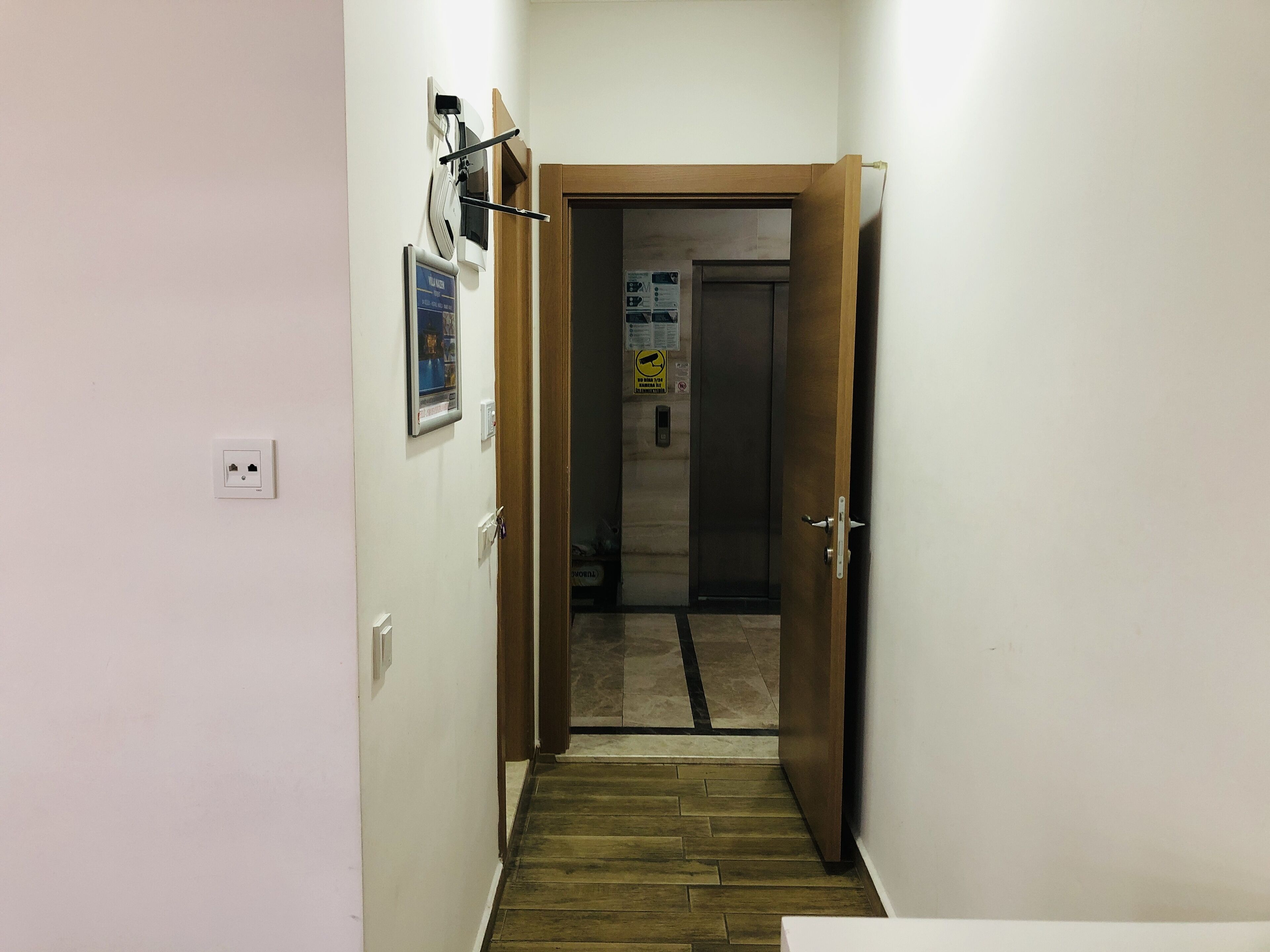 A narrow hallway with a wooden door at the end, a wall socket, and a poster.