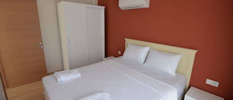 A neatly made bed with white linens and a folded towel, a white nightstand, a white wardrobe, and a wall-mounted air conditioner in a room with red walls.