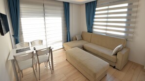 A modern living room with a beige sofa, a dining table with chairs, and large windows with blinds. - Artim Exclusive Apart Hotel (Fethiye)