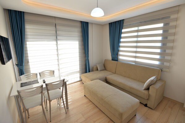 A modern living room with a beige sofa, a dining table with chairs, and large windows with blinds. - Artim Exclusive Apart Hotel (Fethiye)