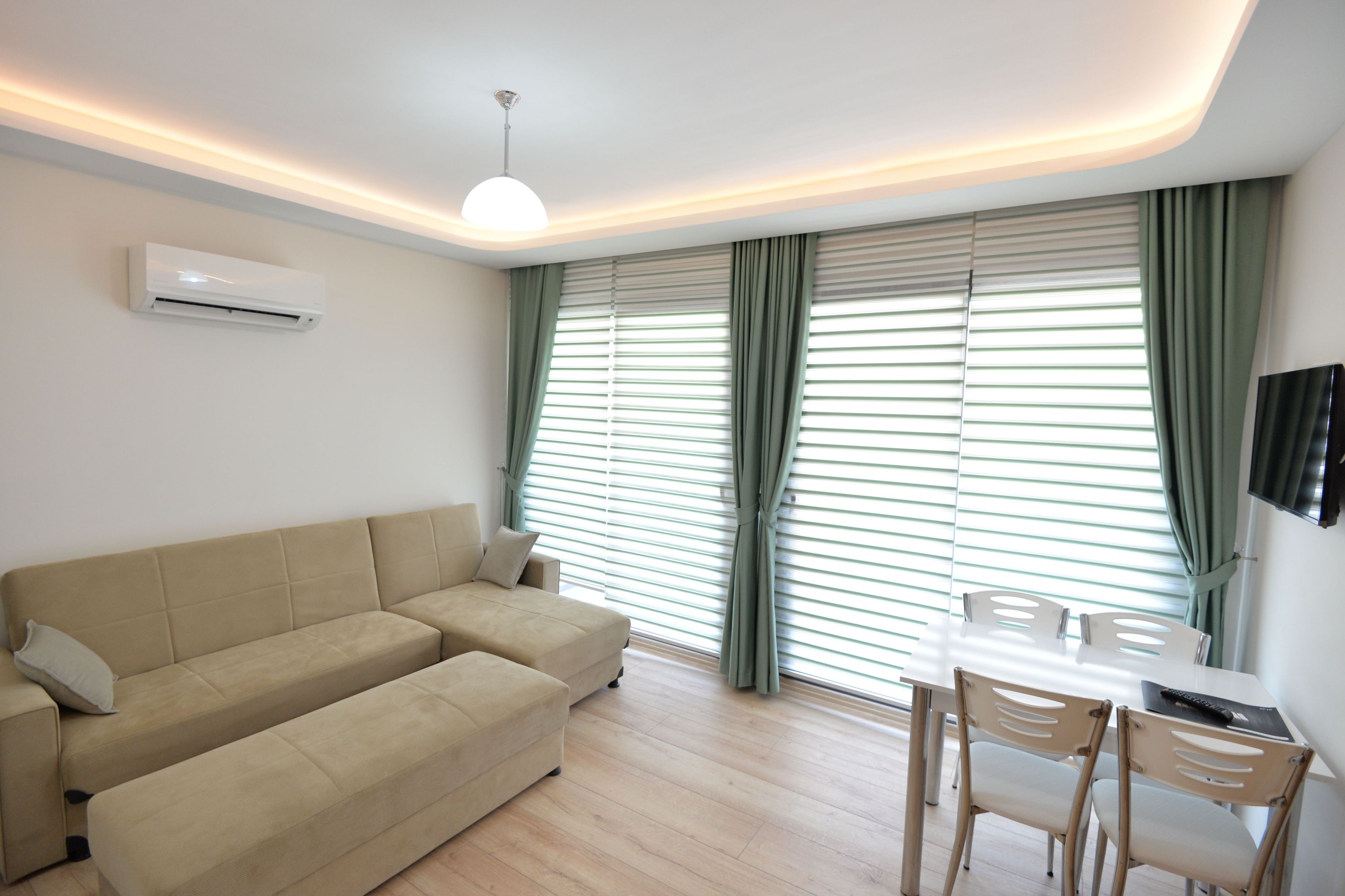 A modern living room with a beige sofa, a dining table with chairs, and a flat-screen TV mounted on the wall.