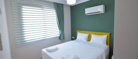 Una cama bien tendida con sábanas y toallas blancas, cabecera amarilla y unidad de aire acondicionado blanca sobre una pared verde.
