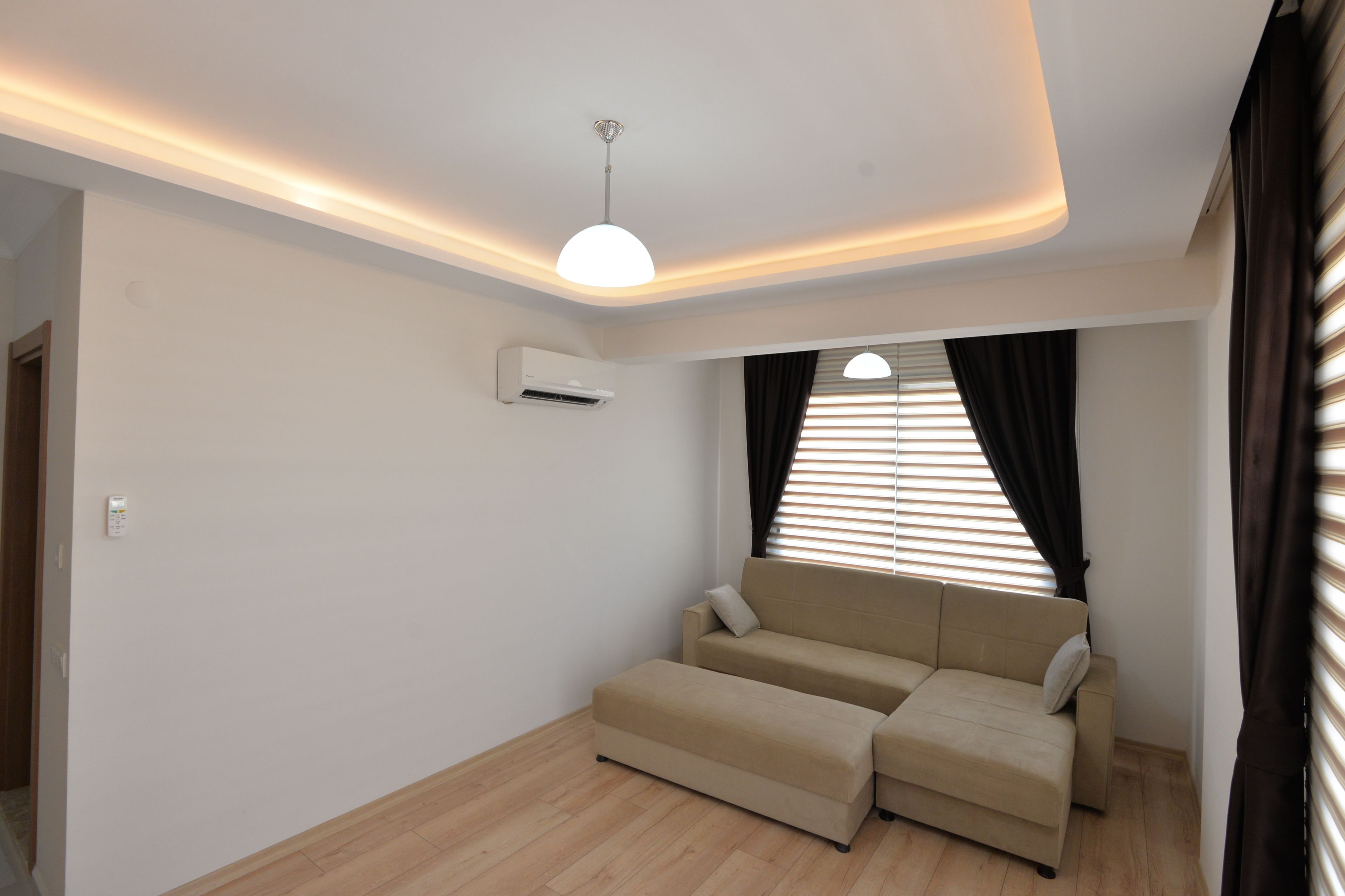 Un salon moderne avec un canapé beige, un luminaire au plafond et une fenêtre équipée de stores.