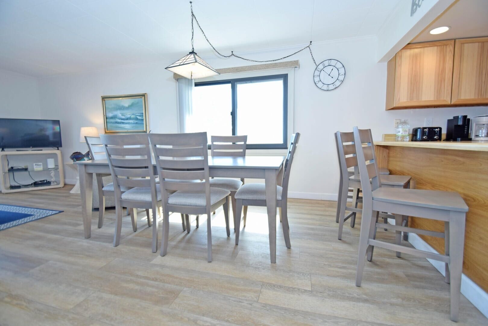 Een moderne keuken met houten kastjes, een eethoek met tafel en stoelen, een televisie en een groot raam.