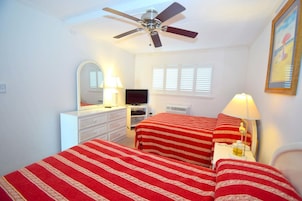 Un dormitorio con dos camas, ventilador de techo, televisor, cómoda y ventana con persianas.