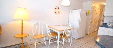 Una cocina pequeña con armarios blancos, un refrigerador blanco, una mesa de madera con cuatro sillas blancas, una lámpara y un flotador en la pared.