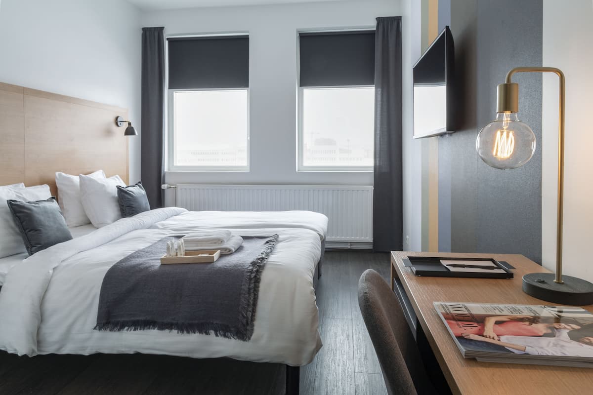 A modern hotel room with a neatly made bed, a side table with a magazine, and a large window offering a view of the city.