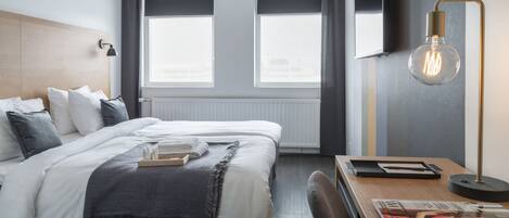 Una habitación de hotel moderna con una cama bien hecha, una mesita de noche con una revista y un amplio ventanal con vistas a la ciudad.