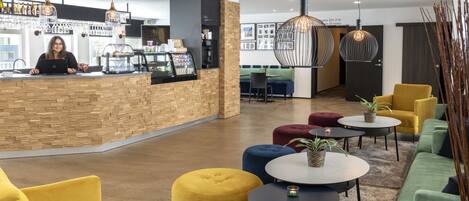 A modern hotel lobby with a wooden reception desk, seating area with colorful ottomans, and a bar with hanging lights.