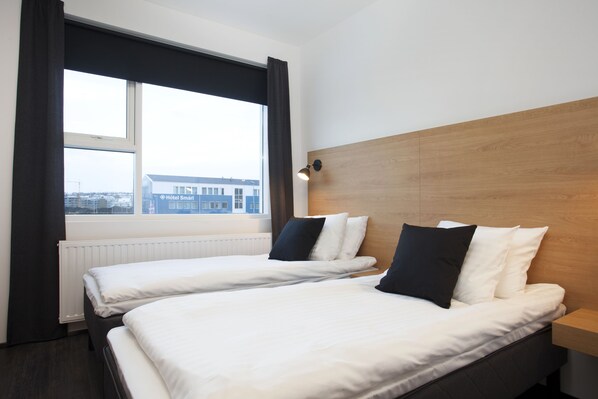 A hotel room with two beds, a wooden headboard, a window with curtains, and a view of buildings outside. - 201 Hotel (Kópavogur)