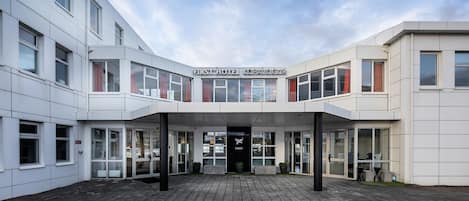 Ein moderner Hotel Eingang mit blauem Himmel, einem Schild mit der Aufschrift „FIRST HOTEL KOPAVOGUR“ und einem Doppeltür-Eingang.