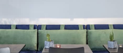 Eine Sitzecke mit einem blauen Sofa, grünen Kissen, einem Holzregal mit Pflanzen und einem Tisch mit Stühlen.