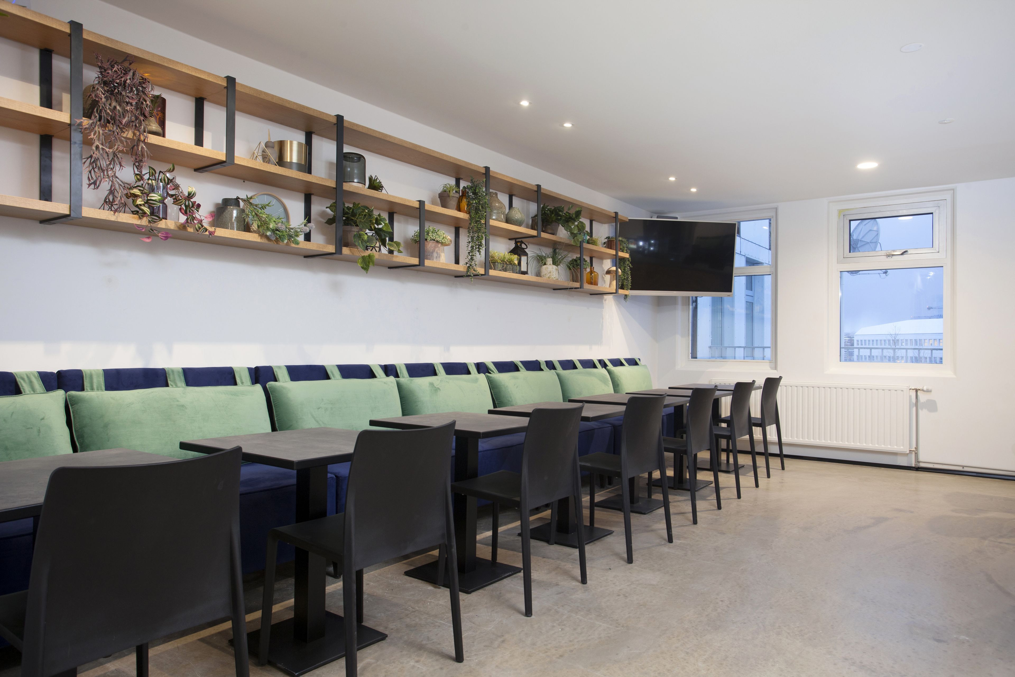 A modern dining area with a long bench, multiple tables, and chairs. There are shelves with plants and decorative items on the wall, and a flat-screen TV mounted above the bench.