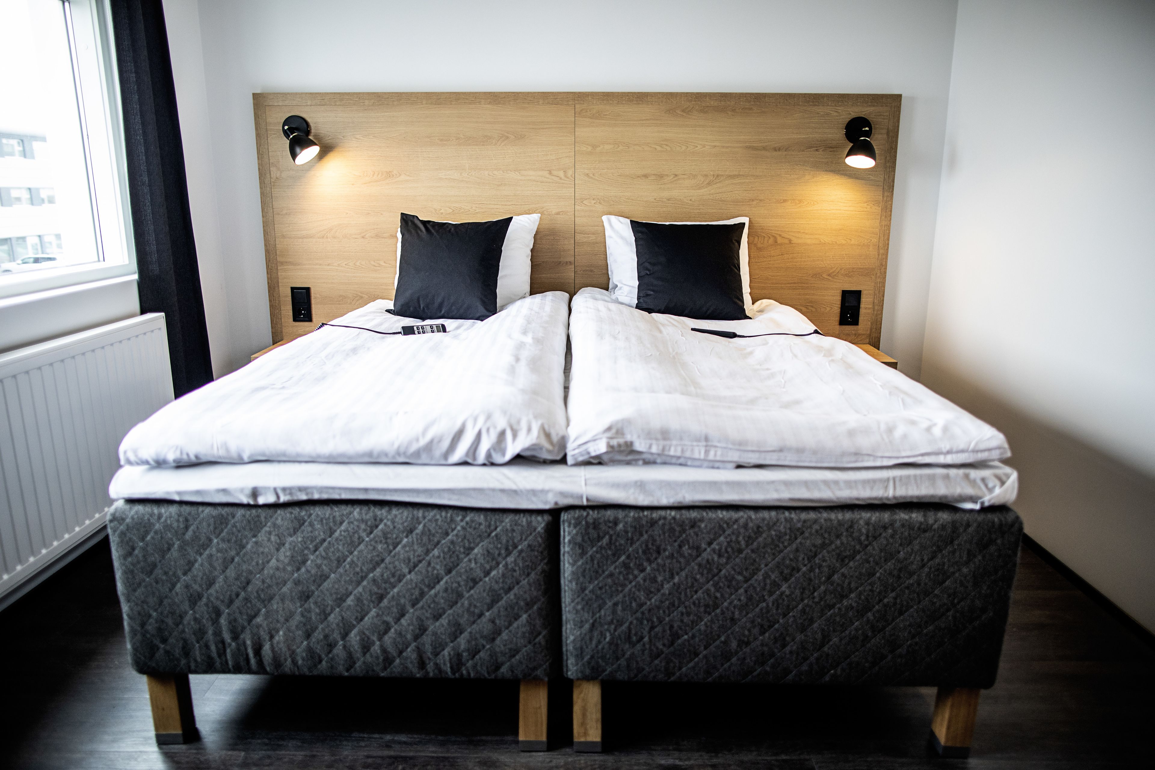 A double bed with a wooden headboard, two bedside lamps, and a view of the city through a window.